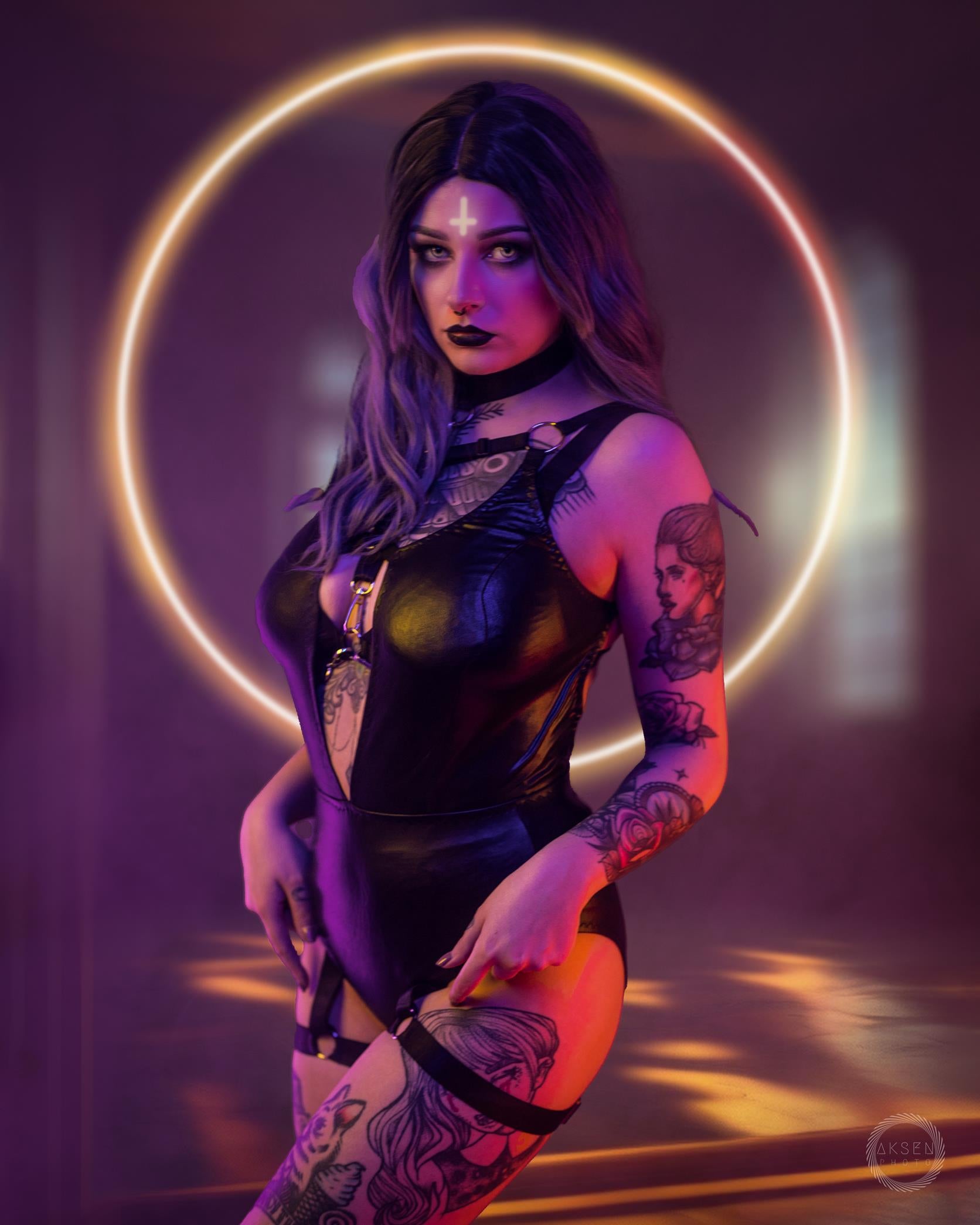 Woman in a black bodysuit with tattoos standing in front of a glowing circular light.