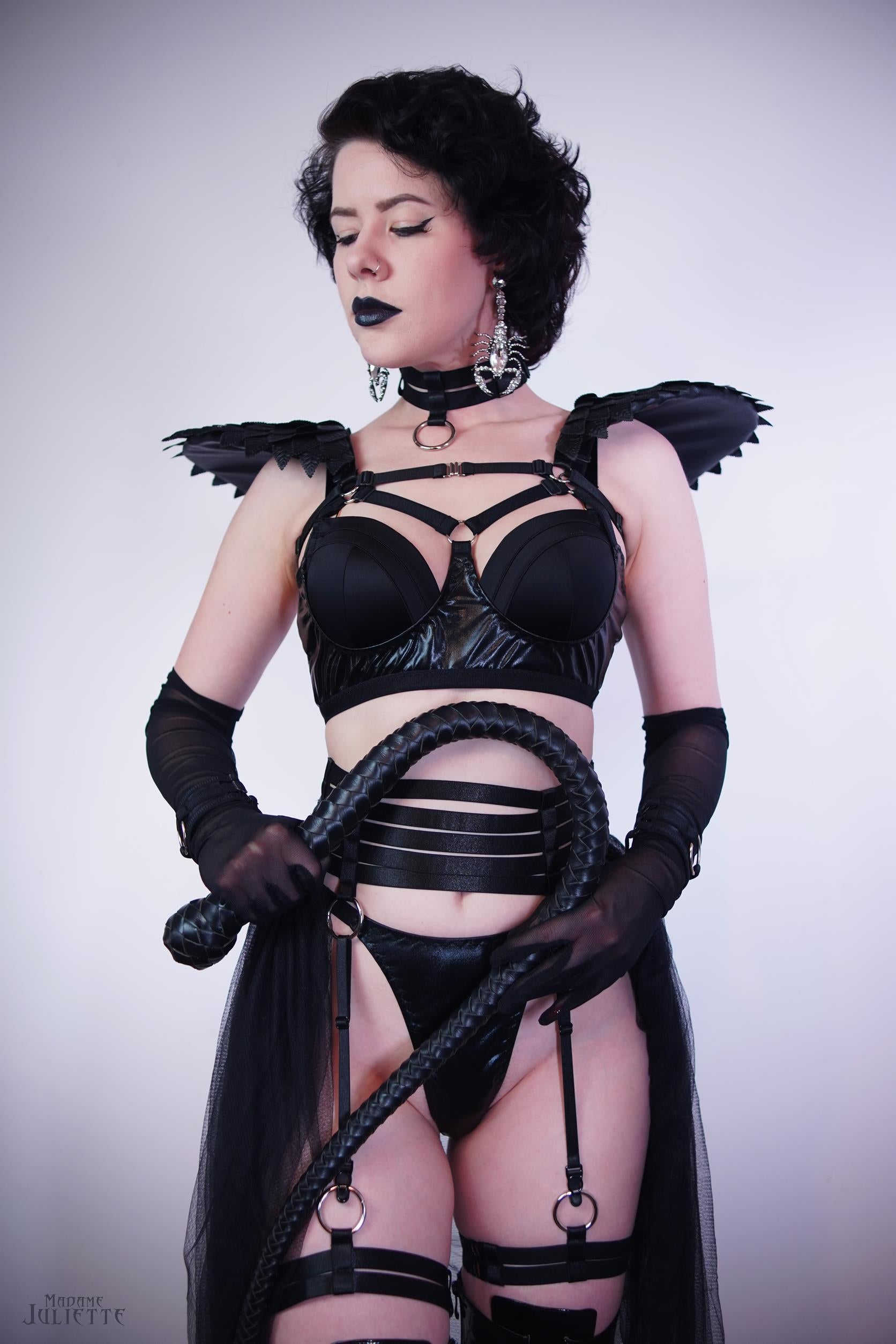 Person wearing a black satin shine strappy garter belt with wings and a whip on a plain background