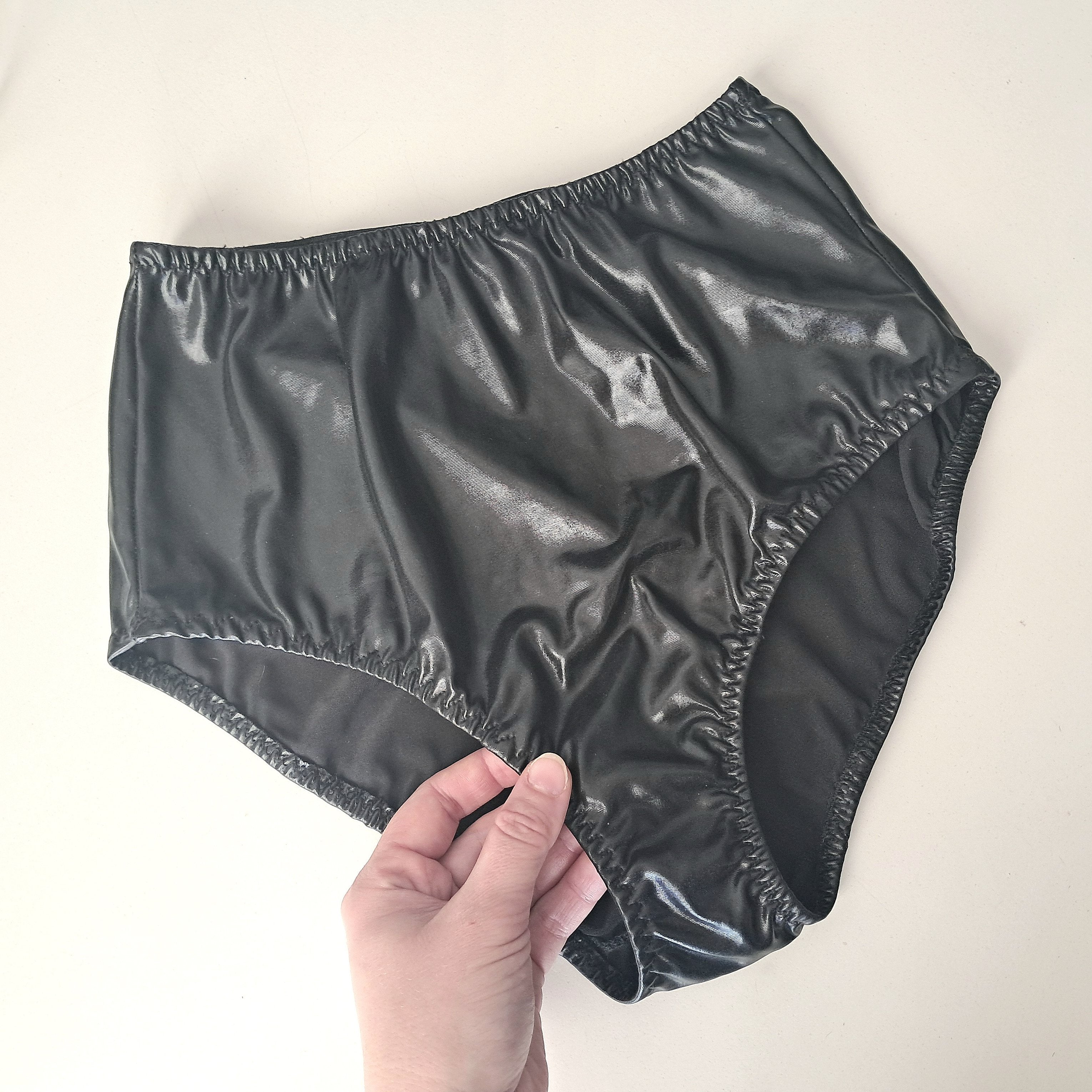 Shiny black wet-look high waist panties held by a hand on a light background