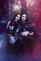 Two women in dark, gothic-style clothing and wet-look high waist panties standing in a forest setting with a mystical atmosphere.