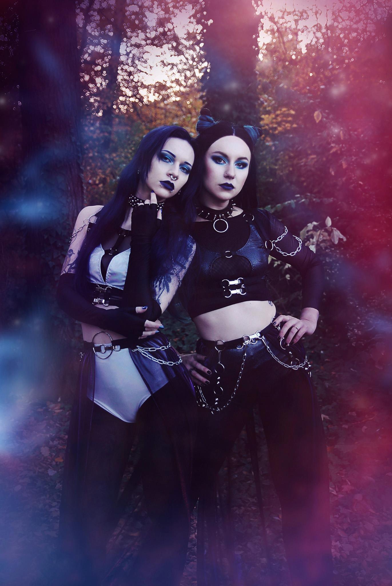 Two women in dark, gothic-style clothing and wet-look high waist panties standing in a forest setting with a mystical atmosphere.