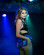 Woman in a black top and blue wet-look open front mini skirt posing on a dark stage with blue lighting.