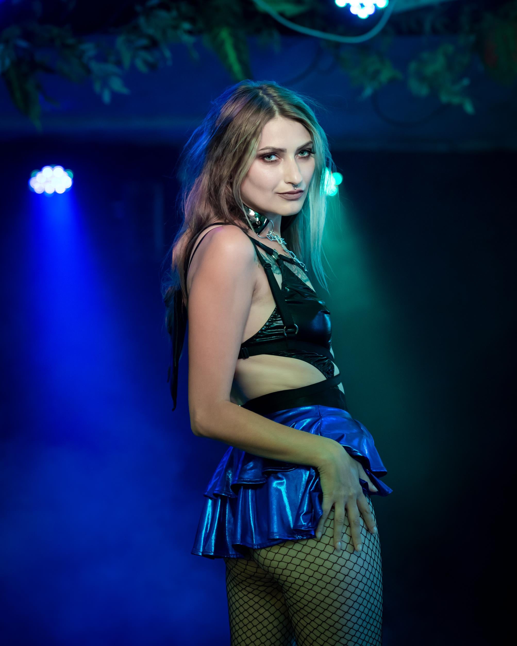 Woman in a black top and blue wet-look open front mini skirt posing on a dark stage with blue lighting.