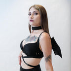 Woman wearing a black harness with bat wings with straps and a choker, standing against a plain background.