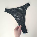Black wet look high waist thong held by a hand on a white background