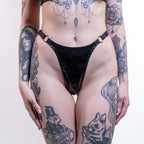 Person wearing a black strappy wet look thong with tattoos on arms and legs against a white background