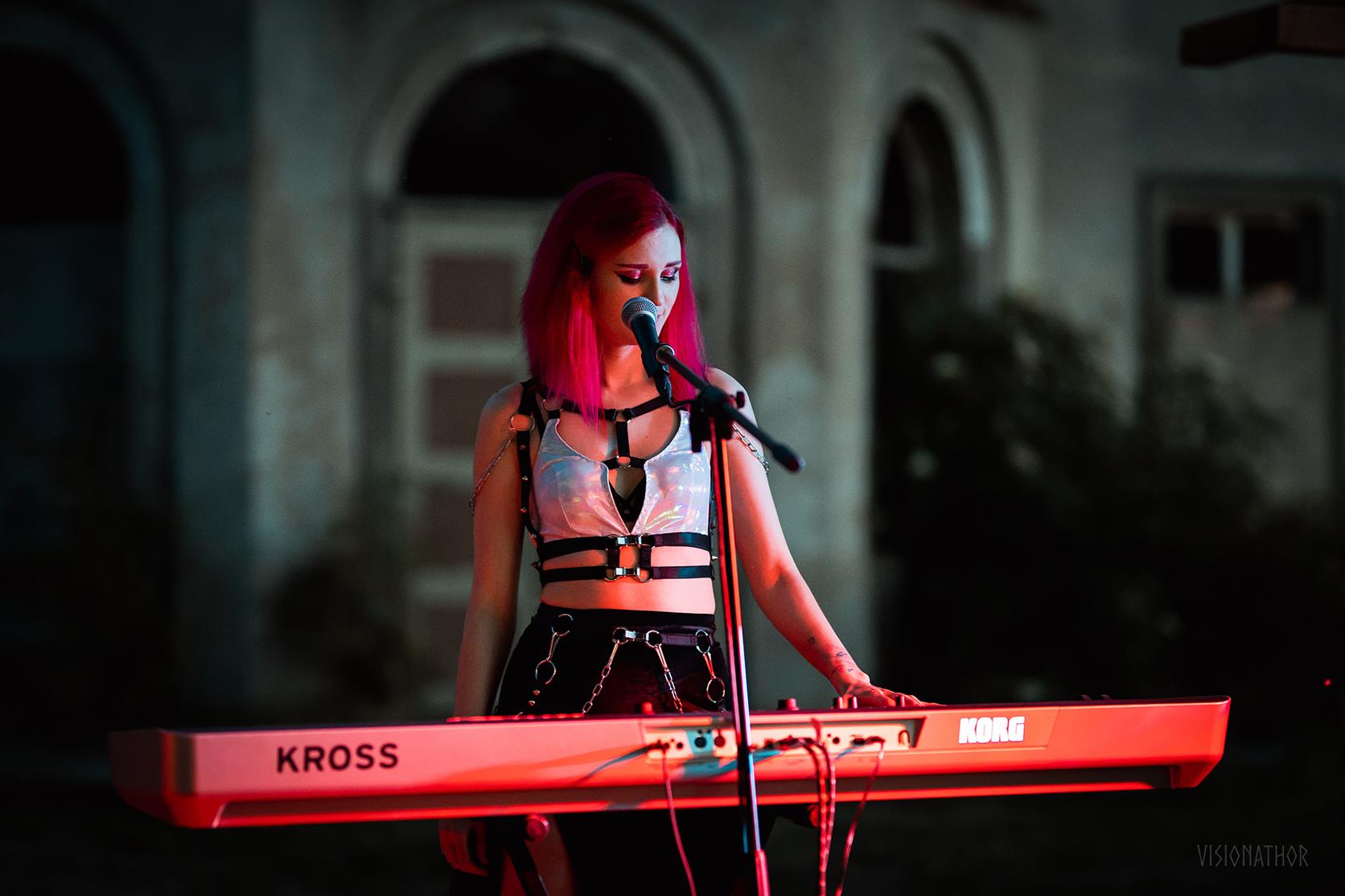 Person with red hair wearing wet look bra playing a KROSS keyboard in a dimly lit setting