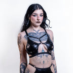 Woman wearing a black cut-out wet look bra with intricate designs on a plain background