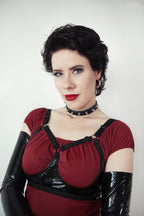 Woman wearing a red top with black straps, open cup wet look bra and black gloves against a white background