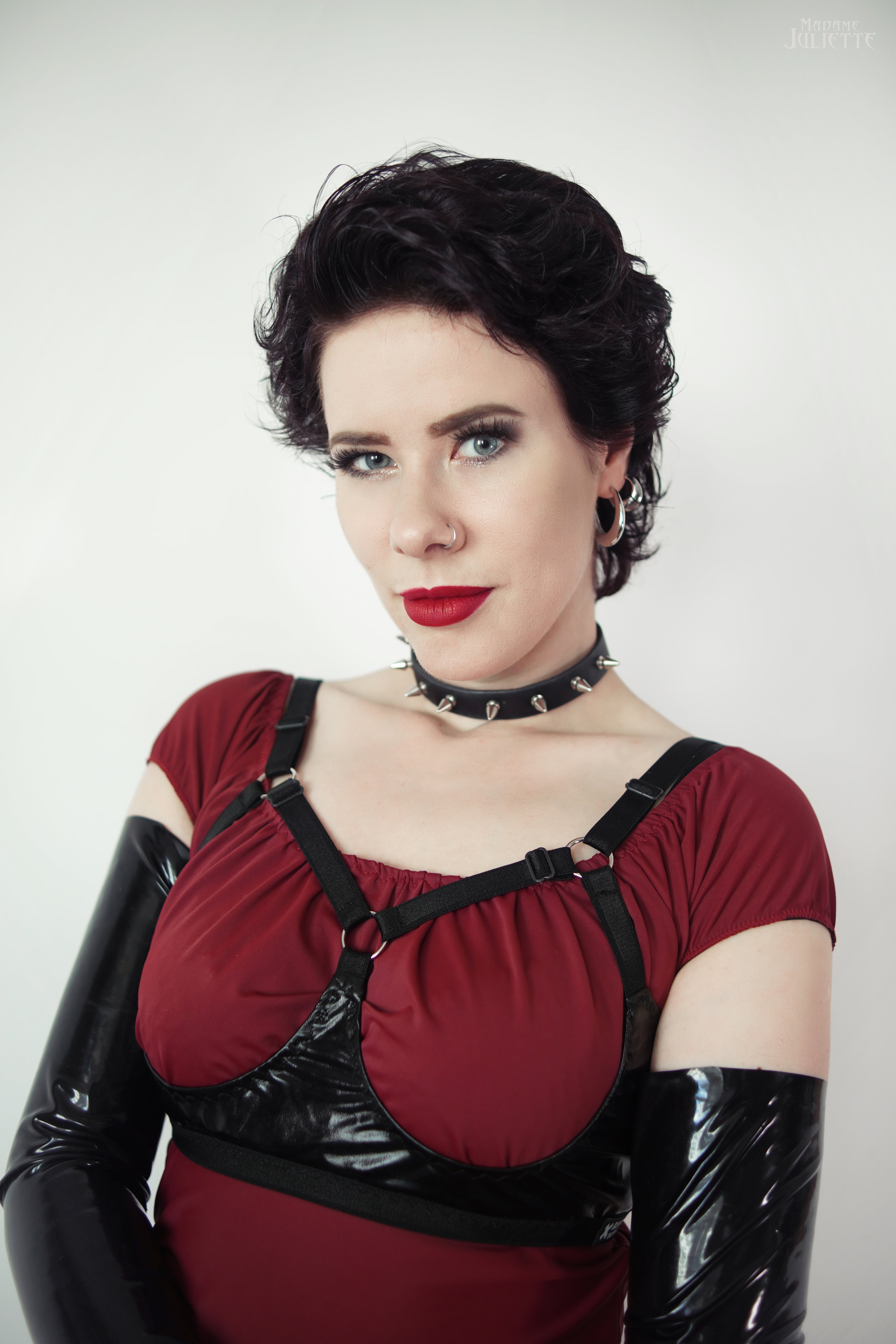 Woman wearing a red top with black straps, open cup wet look bra and black gloves against a white background