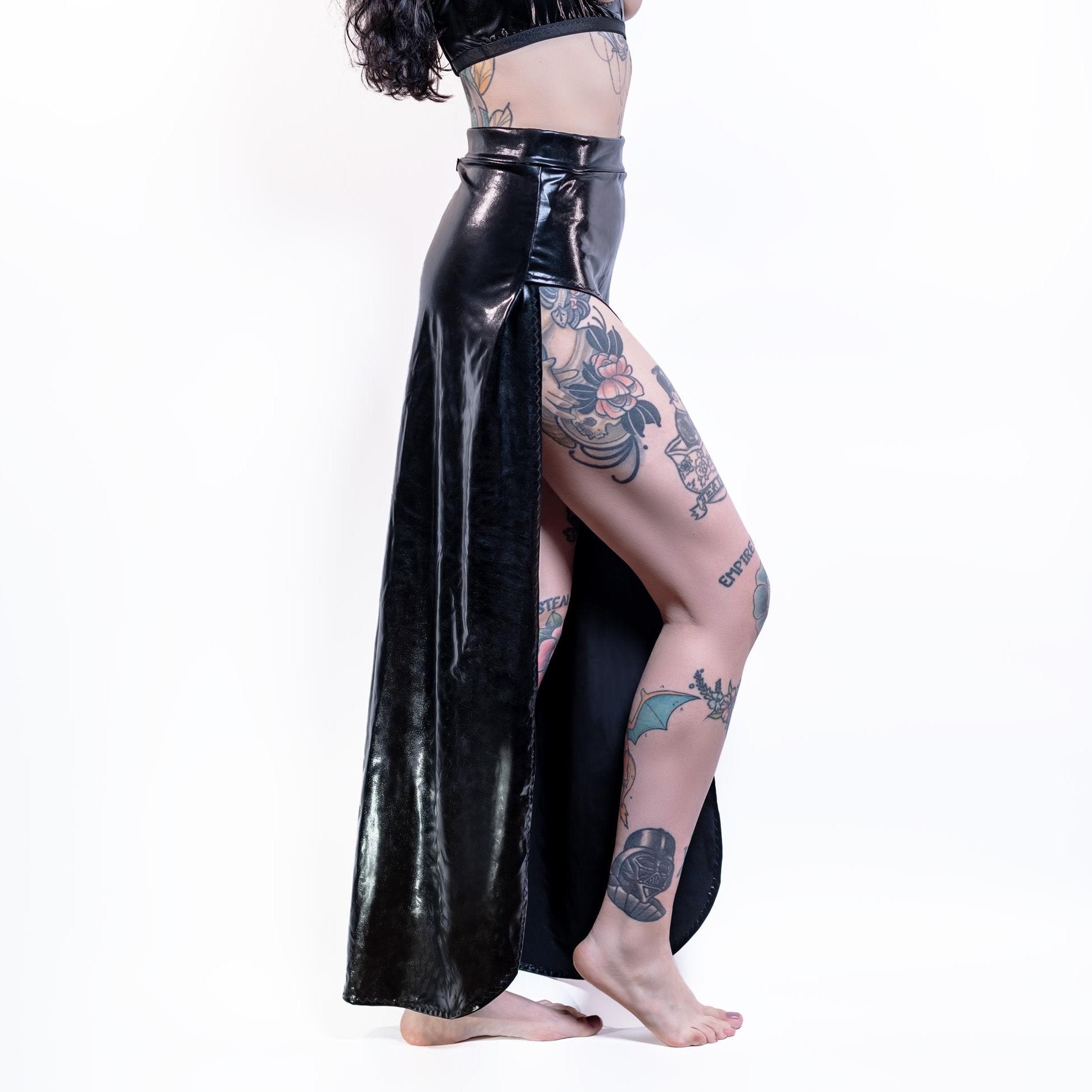 Person wearing a black wet-look cut-out maxi skirt with tattoos visible on the legs against a white background