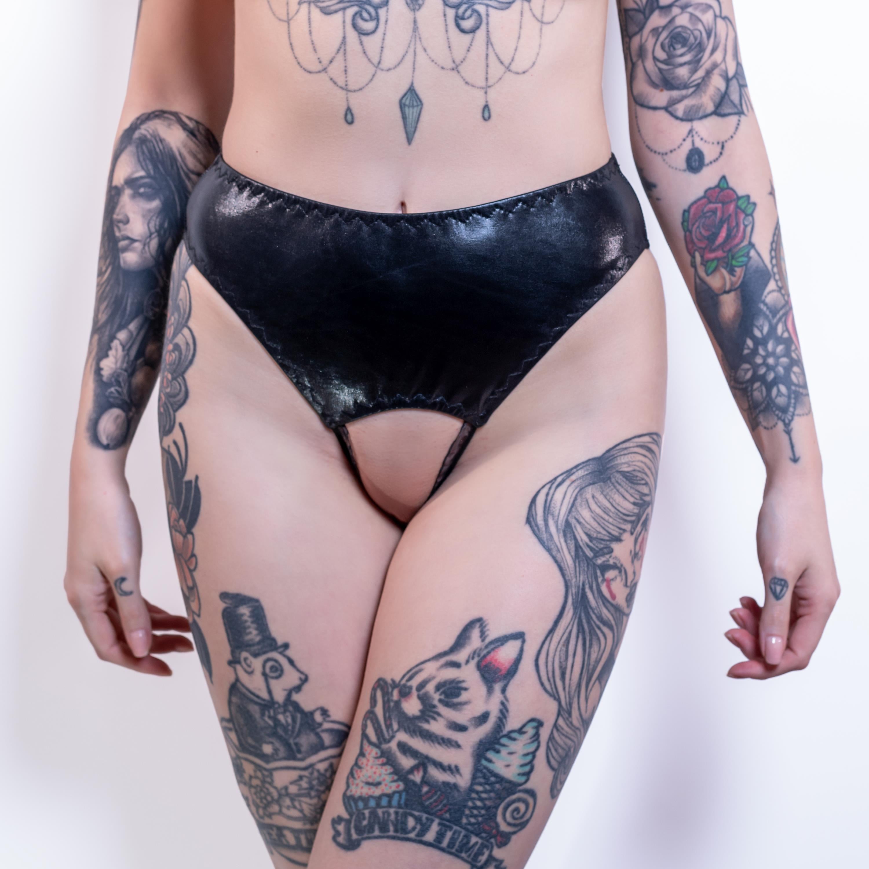 Person wearing black wet-look open crotch panties with tattoos on arms and legs against a white background