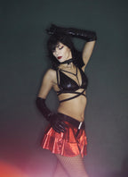 Woman in a black wet look triangle bra and red metallic skirt with black gloves against a dark background