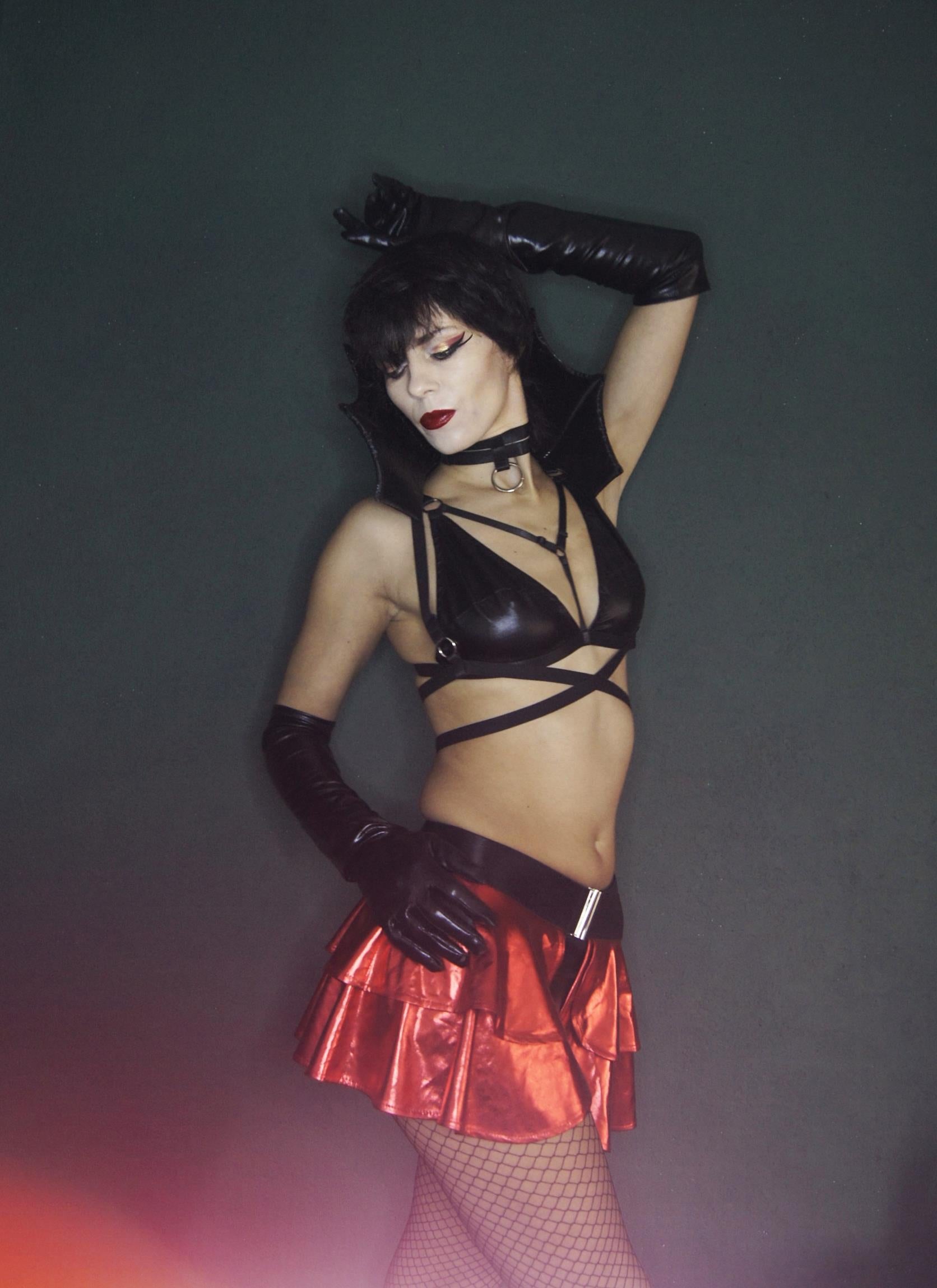 Woman in a black wet look triangle bra and red metallic skirt with black gloves against a dark background