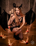 Person in a black wet look cut out dress with wings and horns, surrounded by candles on a wooden floor.