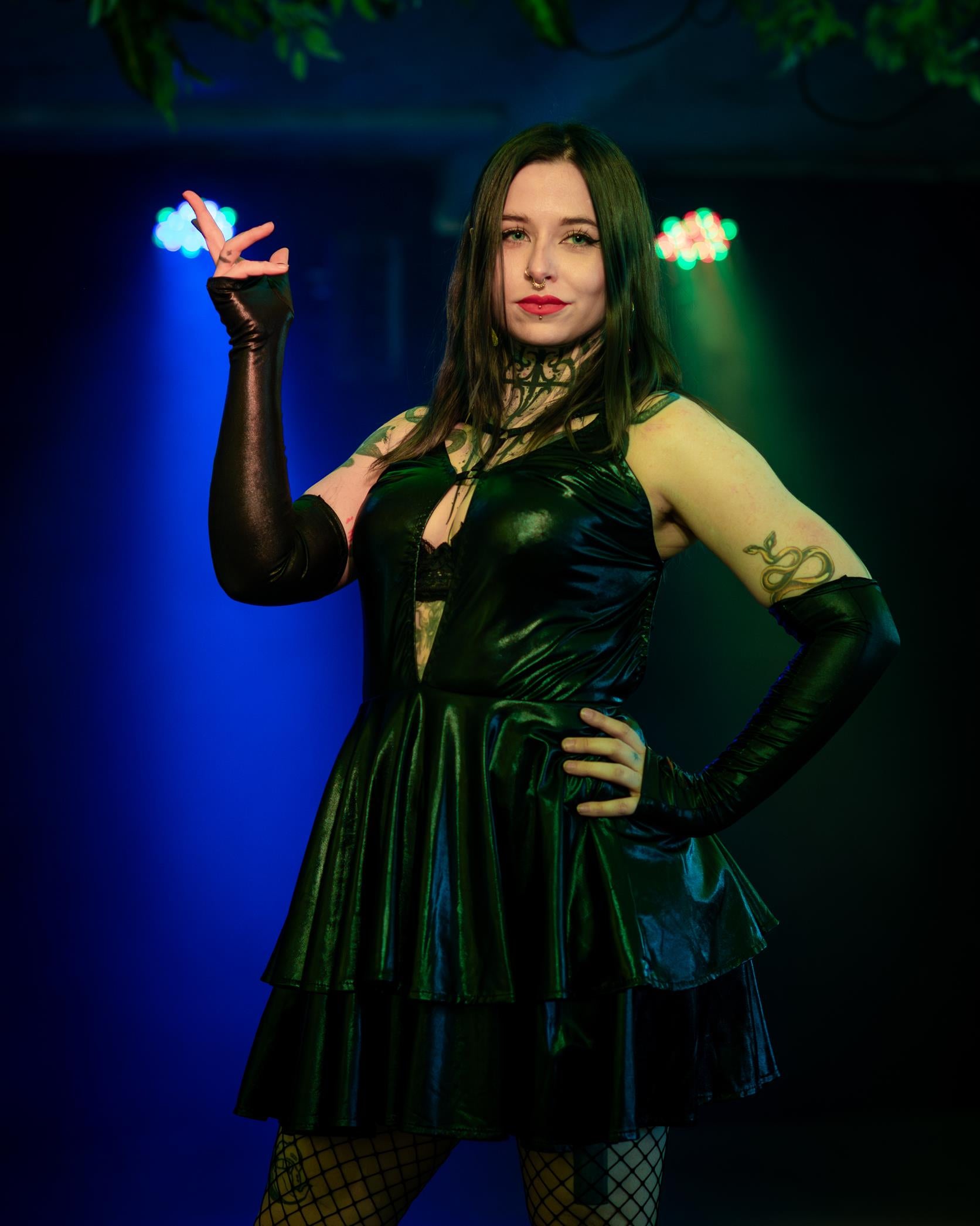 Woman in a wet look flared dress with tattoos posing against a dark background with blue and green lights.