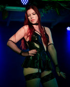 Woman in a black wet look ruffle dress with suspenders with fishnet stockings posing against a dark background