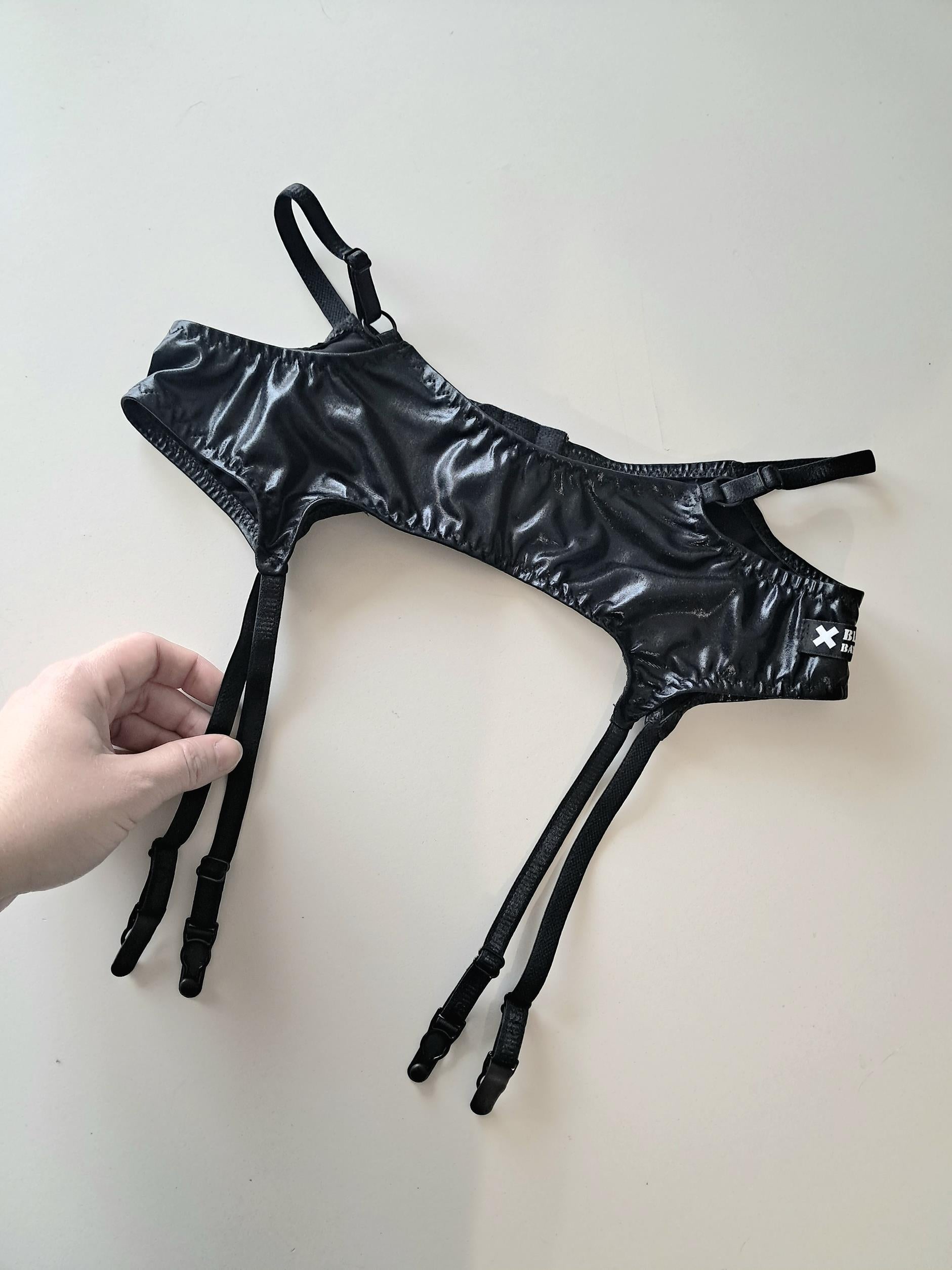 Black wet look strappy garter belt held by a hand on a light gray background